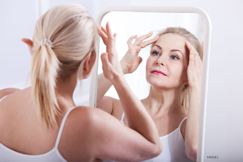 Middle-aged woman looking at herself in the mirror as she pulls her facial skin gently back