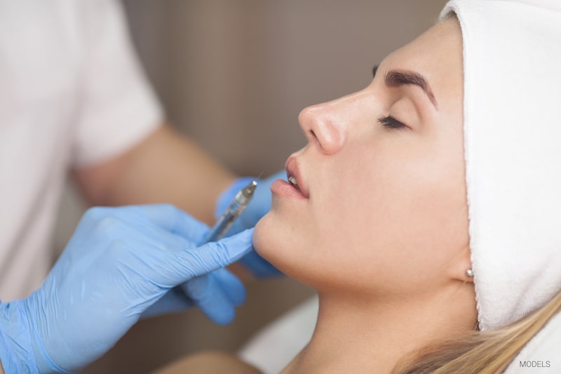 Woman lying back with eyes closed while medical professional prepares to inject a dermal filler.